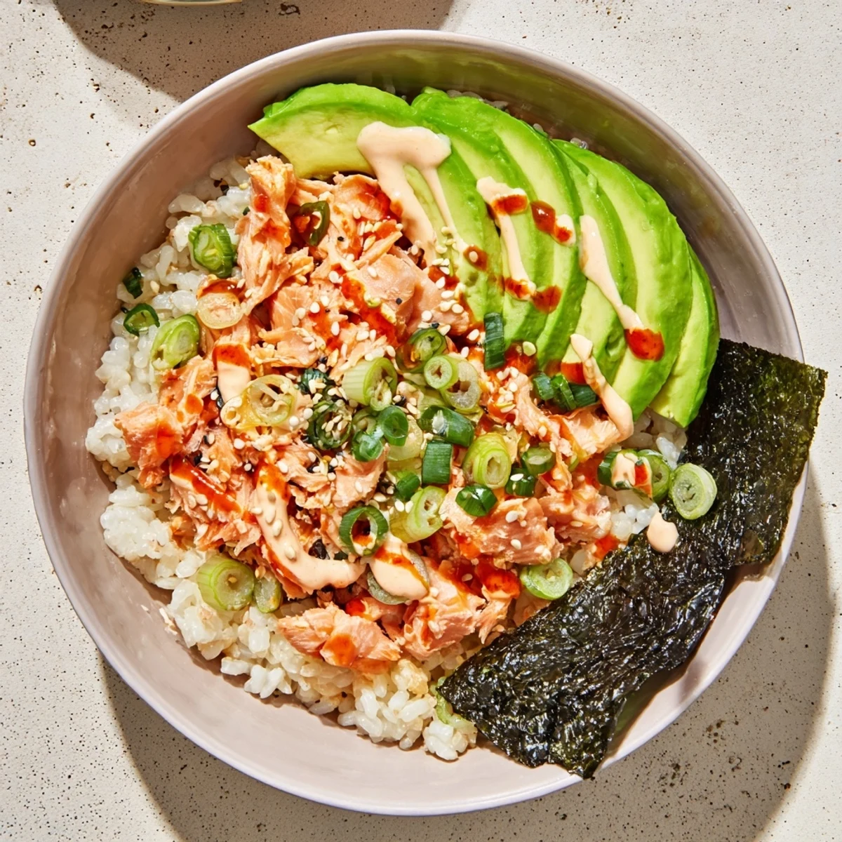 Close-up of a freshly assembled Emily Mariko Salmon Rice Bowl with flaked salmon, creamy avocado slices, and spicy mayo drizzle.
