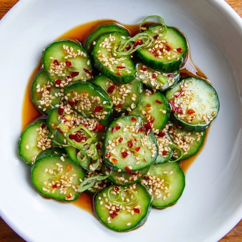 Vibrant green cucumber slices glistening with soy-sesame glaze, garnished with sesame seeds and scallions in a bowl.