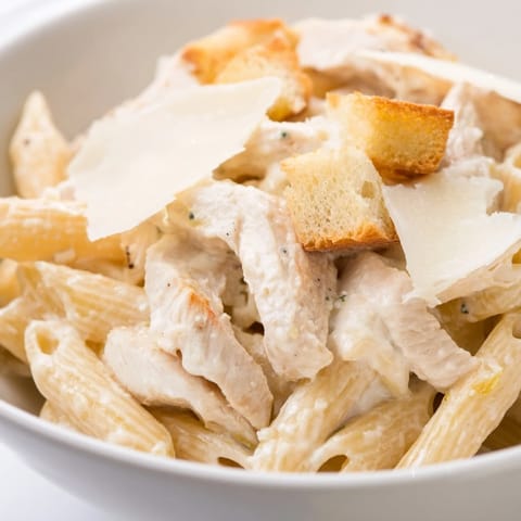 Freshly grated Parmesan and garlic croutons top this vibrant Caesar Pasta Chicken Bowl, served in a rustic white bowl.  