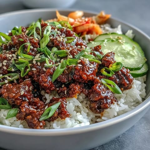 A close-up of a Korean Beef Bowl, featuring glossy gochujang beef on fluffy white rice, topped with quick-pickled carrots and radish, fresh cucumber slices, and spicy kimchi.